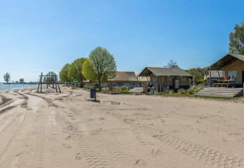 Safari tents at Zuiderzee beach, Netherlands, with trees, playground, clear blue sky, and calm water.