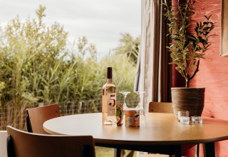 Ronde tafel in een lodge met wijnfles, twee glazen, Pringles en plant naast een raam met uitzicht op groen.