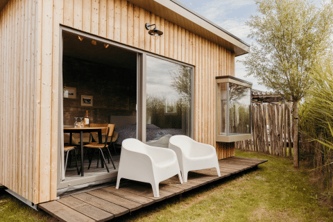 Buitenfoto van een moderne houten lodge met twee witte stoelen op het terras en grote glazen schuifdeur.
