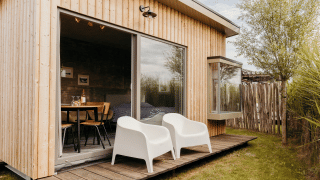 Photo extérieure d’un lodge moderne en bois avec deux chaises blanches sur la terrasse et grandes baies vitrées.