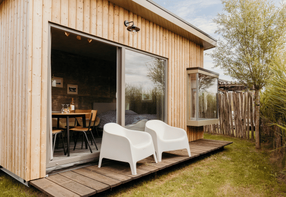 Foto exterior de un lodge moderno de madera con dos sillas blancas en la terraza y grandes ventanales.