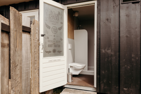 Afbeelding van een lodgebadkamer met open deur, hangtoilet en houten wandpanelen buiten.