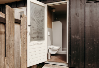 Afbeelding van een lodgebadkamer met open deur, hangtoilet en houten wandpanelen buiten.