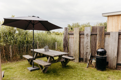 Área de comedor al aire libre en un lodge con mesa de madera, sombrilla, barbacoa y valla de madera.