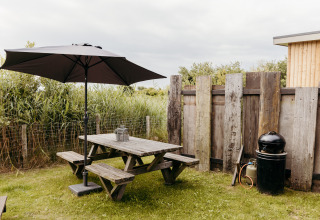 Área de comedor al aire libre en un lodge con mesa de madera, sombrilla, barbacoa y valla de madera.