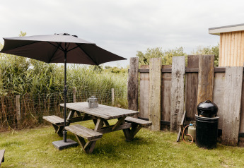 Area esterna di un lodge con panchina in legno, grande ombrellone, barbecue e recinzione rustica.