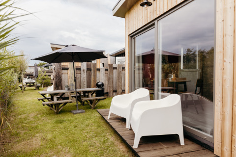 Buitenruimte bij een lodge met witte stoelen, picknicktafel, grote parasol en een grasveld.