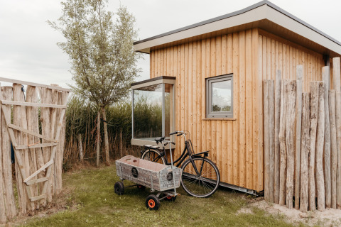 Foto van een houten lodge-hut met fiets en karretje buiten, omgeven door een rustiek houten hek.
