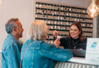 Una recepcionista sonriente entrega llaves a dos huéspedes durante el check-in en un alojamiento glamping.