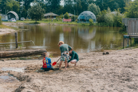 Kinderen spelen in het zand aan een meer op een glampinglocatie met koepeltenten en bomen op de achtergrond.