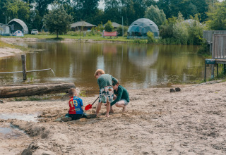 Bambini giocano nella sabbia vicino a un lago in un campeggio glamping con tende a cupola e alberi dietro.