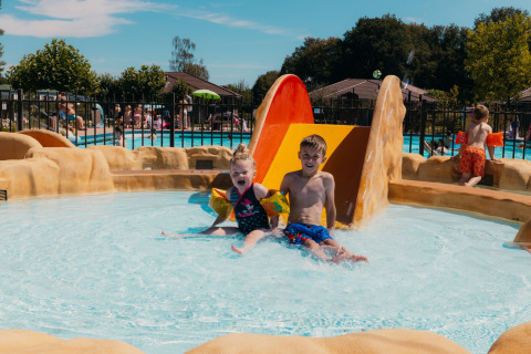 Two kids sliding into a shallow pool area at Holiday park De Boshoek, Gelderland, on a sunny day.