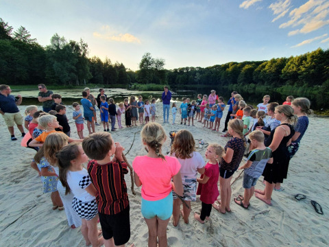 Kinderen en volwassenen staan in een cirkel rond een kampvuur op het strand bij Camping 't Veld.