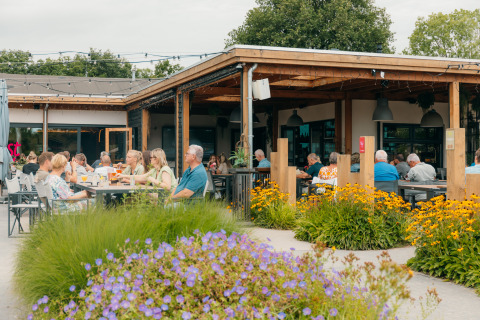Außenbereich des Restaurants im Ferienpark De Boshoek mit Gästen, Blumen und entspannter Atmosphäre.