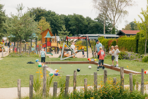 Børn leger udendørs ved en legeplads i en feriepark i Gelderland, Holland, omgivet af grønne planter.