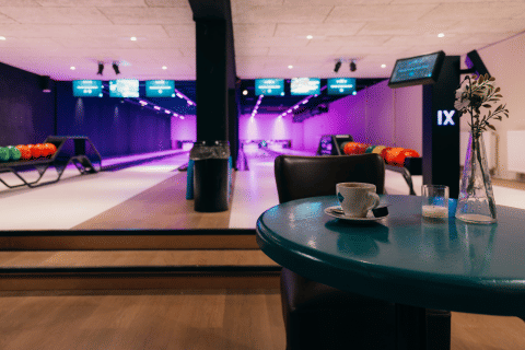 Binnen bowlingbaan met gekleurde ballen, scoreborden en een kopje koffie op een blauwe tafel.