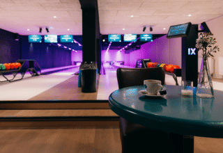 Salle de bowling intérieure avec boules colorées, tableaux d’affichage et tasse de café sur table bleue.