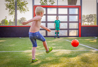 Deux enfants jouent au football sur un terrain dans un parc vacances glamping, l’un tire, l’autre est gardien.