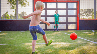 Dos niños juegan al fútbol en un campo de un parque vacacional con glamping, uno chuta y otro es portero.