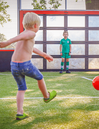 Dos niños juegan al fútbol en un campo de un parque vacacional con glamping, uno chuta y otro es portero.