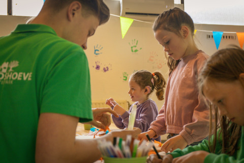 Children and an adult do indoor crafts at a holiday park, with colorful handprints decorating the wall.