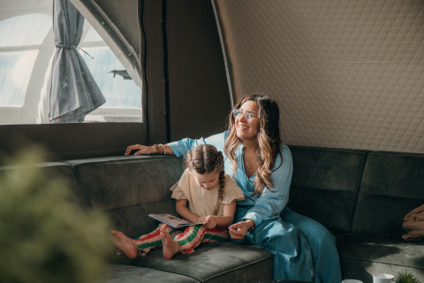 Mère et enfant se détendent dans une tente de glamping au parc de vacances, l'enfant lit sur le canapé.