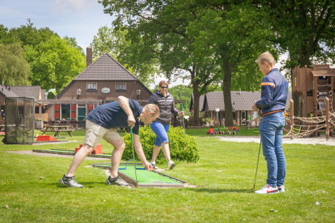 Trois adultes jouent au mini-golf dans un parc de vacances avec hébergements glamping et aire de jeux.