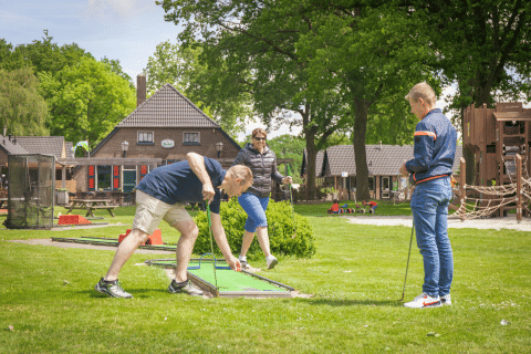 Three adults play mini golf at a holiday park with glamping accommodations and children's play area.