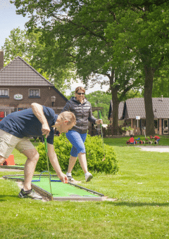 Tres adultos juegan mini golf en un parque vacacional con alojamientos glamping y zona de juegos infantil.