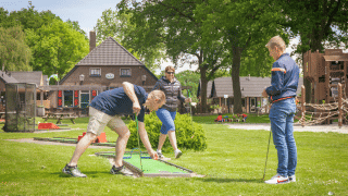 Tres adultos juegan mini golf en un parque vacacional con alojamientos glamping y zona de juegos infantil.