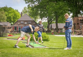 Trois adultes jouent au mini-golf dans un parc de vacances avec hébergements glamping et aire de jeux.