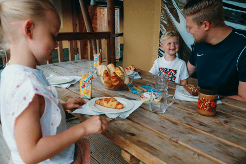 Familie geniet van ontbijt met croissants en fruitsap op het terras van een glampingverblijf in een vakantiepark.