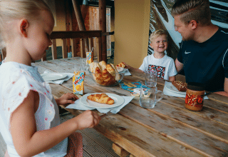 Gezin geniet van ontbijt met croissantjes en sap op het terras bij een glampingaccommodatie in een vakantiepark.