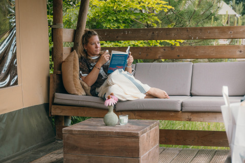 Femme détendue lisant un livre sur un canapé extérieur confortable dans un site de glamping entouré de nature.