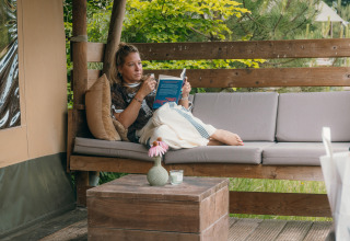 Femme détendue lisant un livre sur un canapé extérieur confortable dans un site de glamping entouré de nature.