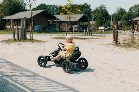 Jongen rijdt met een trapauto op een zandweg voor glampingtenten in een vakantiepark in de zon.