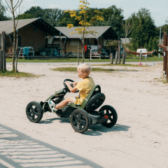 Niño conduce un kart de pedales por un sendero de arena frente a tiendas glamping en un parque vacacional soleado.