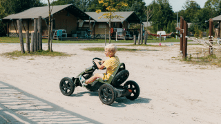 Niño conduce un kart de pedales por un sendero de arena frente a tiendas glamping en un parque vacacional soleado.
