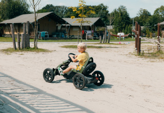 Dreng kører pedalbil på sandsti foran glampingtelte og legeplads i en feriepark under solskin.