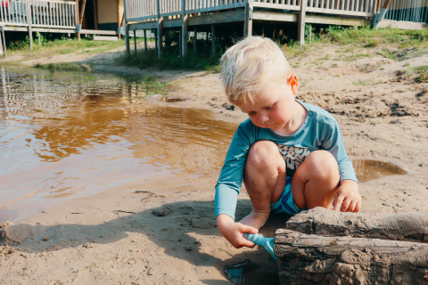 Kleiner Junge spielt am sandigen Ufer eines Teiches beim Ferienpark mit Glamping-Unterkünften im Hintergrund.