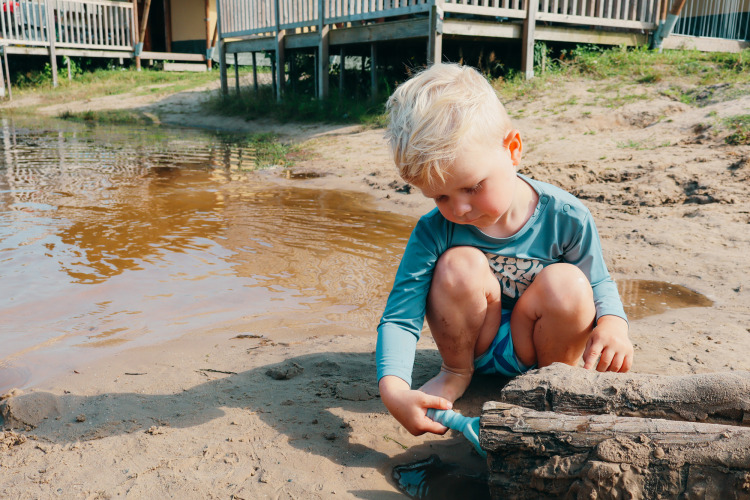 Jonge jongen speelt in het zand bij het water op een vakantiepark met glamping accommodaties op de achtergrond.