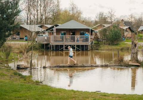 Un enfant traverse un tronc sur un étang devant des lodges glamping dans un parc de vacances en pleine nature.