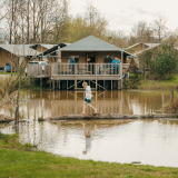 Kind loopt over een boomstam boven vijver bij glampingaccommodaties in een vakantiepark midden in de natuur.