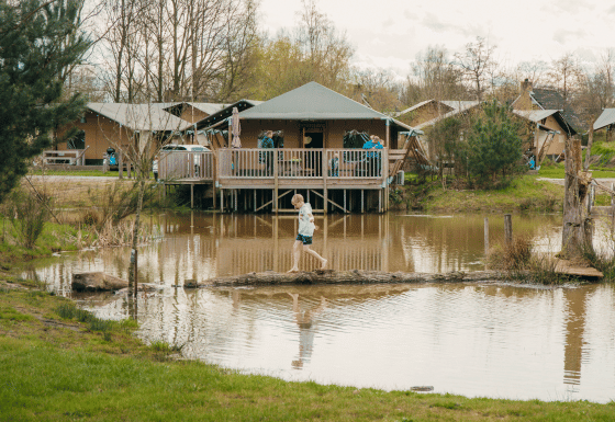 Kind steekt via een boomstam een vijver over bij glampinglodges in een vakantiepark omringd door natuur.