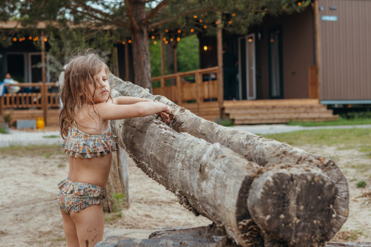 Kleines Mädchen in Badesachen spielt an einem Baumstamm vor Glamping-Hütten in einem Ferienpark.