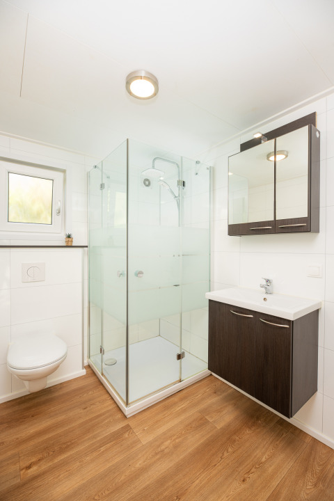 Moderne badkamer met houten vloer, glazen douche, wandtoilet en wastafel in River Cabin met bubbeltub, Holiday Park Mölke.
