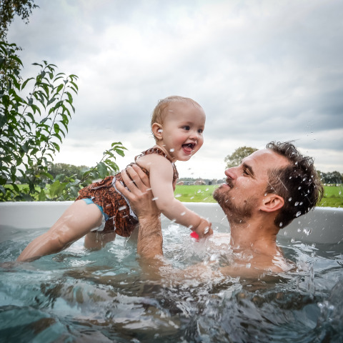 Un padre juega con su bebé en una bañera de hidromasaje al aire libre en River Cabin, Holiday Park Mölke, Países Bajos.