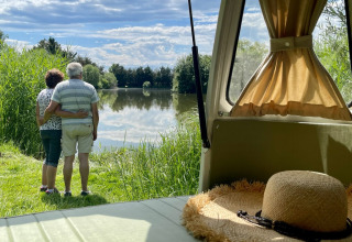 Ældre par omfavner ved en sø, set fra en campingvogn med stråhat, Domaine des Feuilles, Auvergne-Rhône-Alpes.