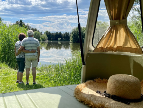 Ældre par omfavner ved en sø, set fra en campingvogn med stråhat, Domaine des Feuilles, Auvergne-Rhône-Alpes.