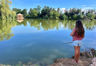 A girl fishing by a tranquil lake with trees and a wooden cabin in the background at Domaine des Feuilles.